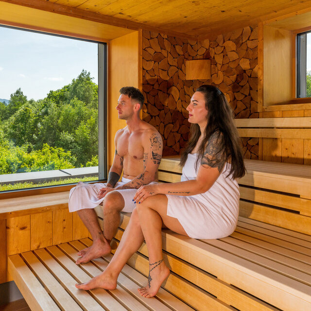 Ein Mann und eine Frau sitzen entspannt auf einer Holzbank in der Panoramasauna - Hotel Herzog Heinrich.
